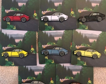 Könnte beinhalten: Satz von zehn Emaille-Pins mit einem Oldtimer-Sportwagen in verschiedenen Farben, darunter Rot, Weiß, Schwarz, Gelb, Blau und Grau. Die Pins sind vor einem Hintergrund aus grünen Bäumen und einer kurvigen Straße angeordnet.