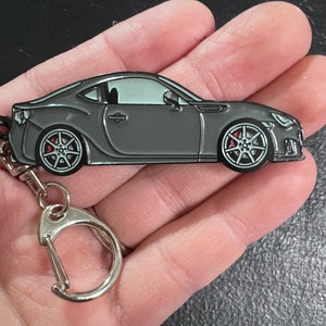 May include: A gray metal keychain featuring a detailed model of a Subaru BRZ sports car. The keychain has a silver metal ring and a silver metal clasp.