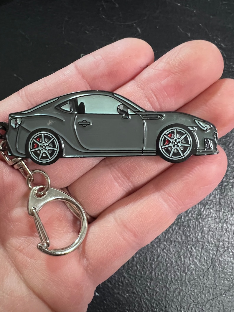 May include: A gray metal keychain featuring a detailed model of a Subaru BRZ sports car. The keychain has a silver metal ring and a silver metal clasp.