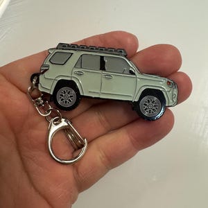 Puede incluir: Un llavero de metal verde claro con forma de Toyota 4Runner con una baca de techo negra. El llavero tiene un anillo de metal plateado.