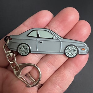 Puede incluir: Un llavero de coche plateado con un anillo de metal plateado. El coche es un coupé plateado con ruedas negras.