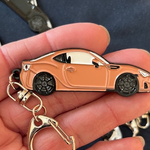 May include: A keychain featuring a small, detailed replica of a light brown sports car. The car has black wheels and a black outline. The keychain is attached to a silver split ring.