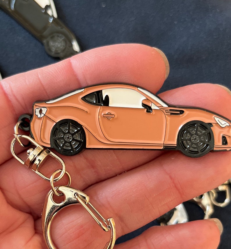 May include: A keychain featuring a small, detailed replica of a light brown sports car. The car has black wheels and a black outline. The keychain is attached to a silver split ring.