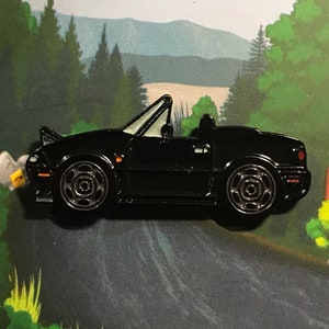 Puede incluir: Pin de esmalte negro de un coche deportivo convertible con interior blanco. El coche está mirando hacia la derecha y tiene un fondo negro.