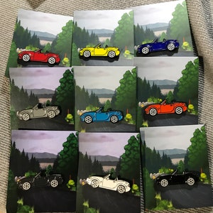 Puede incluir: Un conjunto de diez pines de esmalte que representan diferentes coches Mazda MX-5 de colores. Los pines son todos del mismo tamaño y forma, y todos están colocados sobre un fondo de escena de bosque. Los pines son una excelente manera de mostrar tu amor por el Mazda MX-5.