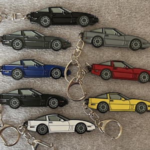 May include: A selection of car keyrings in a range of colours: black, grey, blue, red, yellow, and white. Each keyring has a detailed car design and is attached to a silver-coloured key ring.