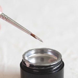 Puede incluir: Un pequeño recipiente plateado con tapa negra, que contiene un gel transparente. Un pincel plateado y blanco se sostiene sobre el recipiente.