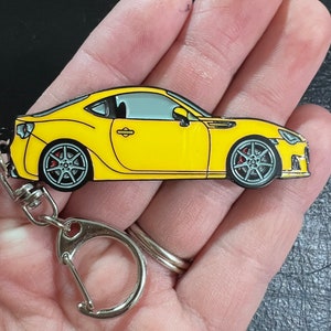 May include: A yellow metal keychain featuring a detailed model of a sports car. The car has black wheels with red brake calipers and a silver windshield. The keychain has a silver metal ring.
