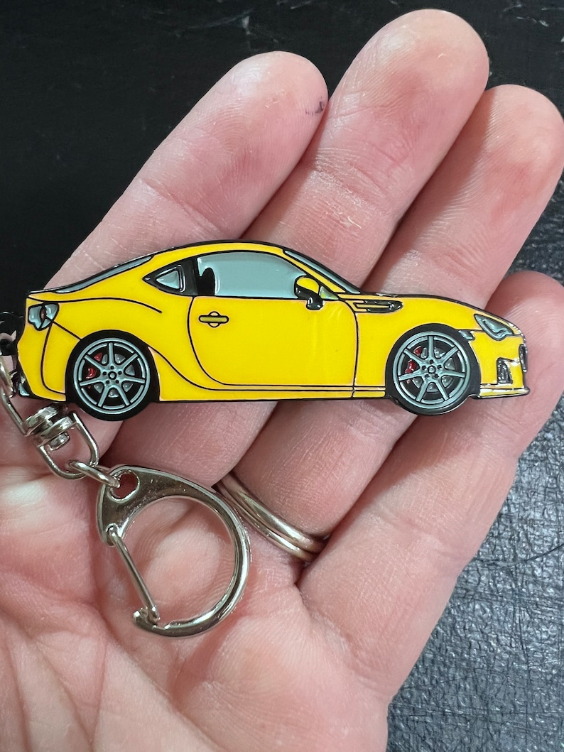 May include: A yellow metal keychain featuring a detailed model of a sports car. The car has black wheels with red brake calipers and a silver windshield. The keychain has a silver metal ring.