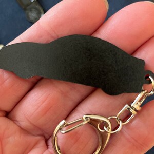 May include: Black car keychain with a silver metal key ring. The keychain is made of a smooth, black material and has a detailed car silhouette.