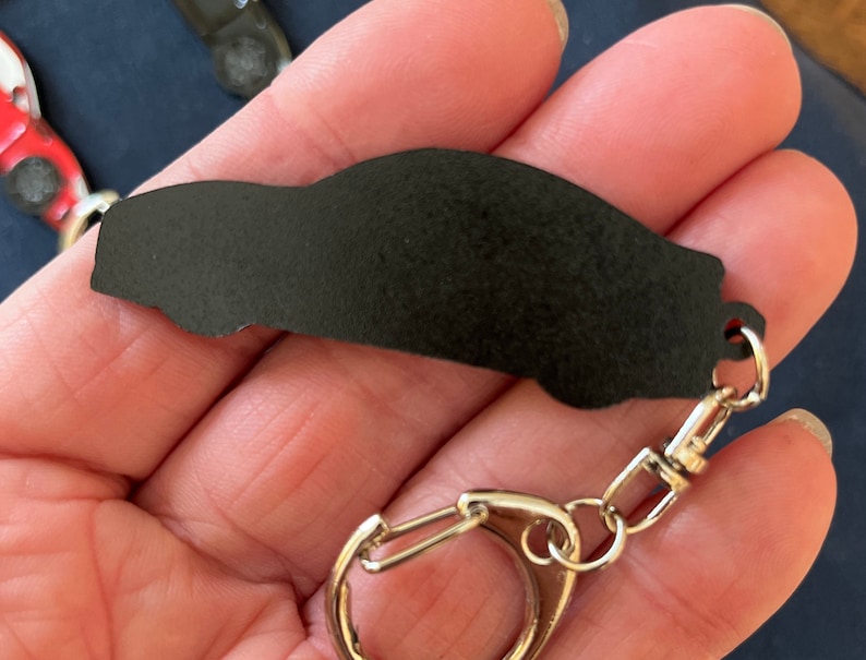 May include: Black car keychain with a silver metal key ring. The keychain is made of a smooth, black material and has a detailed car silhouette.