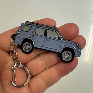 Puede incluir: Un llavero de metal plateado con un modelo detallado de un Toyota 4Runner gris con un portaequipajes en el techo. El llavero tiene un anillo de metal plateado.