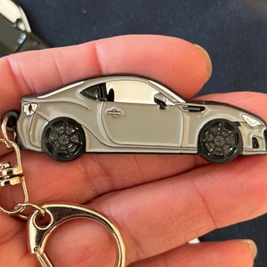 May include: A silver metal keychain featuring a detailed model of a Subaru BRZ sports car. The keychain has a silver metal ring and a black metal clasp.