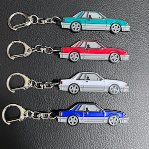 May include: Four keychains featuring a side profile of a vintage car in different colours: teal, red, white, and blue. Each keychain has a silver metal ring.