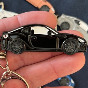 May include: Black metal keychain featuring a detailed model of a sports car. The car has a white roof and black wheels. The keychain has a silver metal ring.