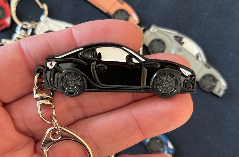 May include: Black metal keychain featuring a detailed model of a sports car. The car has a white roof and black wheels. The keychain has a silver metal ring.