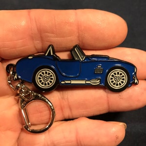 May include: A blue metal keychain shaped like a classic sports car. The car has a silver grille and white wheels. The keychain has a silver metal ring.