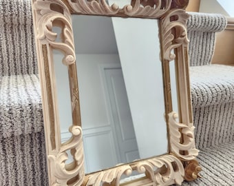 May include: A rectangular mirror featuring an ornate, cream-colored frame with intricate carved scrollwork and leaf motifs. The inner edges of the frame are accented with metallic gold detailing, providing a contrast to the light-colored exterior.