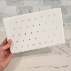 May include: A rectangular white box with a textured surface featuring a repeating pattern of small gold fleur-de-lis symbols across the front. The box has rounded corners and a clean, simple design, presenting a consistent aesthetic throughout.