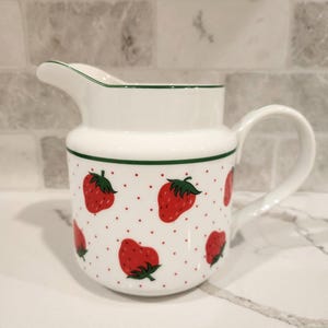 May include: White ceramic pitcher with a handle and spout, decorated with red strawberries and small red dots on a white background. The rim and a band around the middle are trimmed with green. A charming addition to any kitchen.