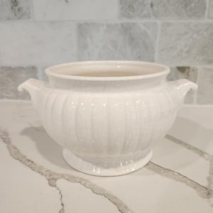 May include: A white ceramic sugar bowl featuring a rounded body with vertical ribbed detailing, a flared rim, and two small side handles. The surface has a subtle crackle glaze finish and sits on a circular pedestal base.