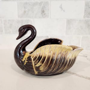 May include: A ceramic swan-shaped bowl featuring a glossy dark brown glaze on the neck and base, transitioning into a textured cream and yellow drip glaze across the wing area. The piece has a curved neck and a hollow, open center body.