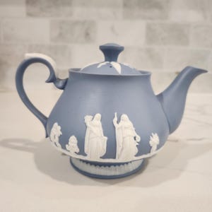 May include: A light blue teapot with a matte finish featuring white relief figures of classical individuals and floral motifs. The teapot includes a matching lid with a central knob, a curved handle, and a spout, all decorated with white accents.
