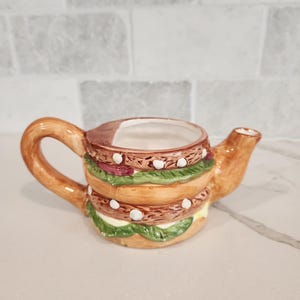 May include: A novelty ceramic teapot shaped like a hamburger. The teapot features a brown bun, lettuce, tomato, and a white interior. The handle and spout are brown. A unique kitchen accessory.