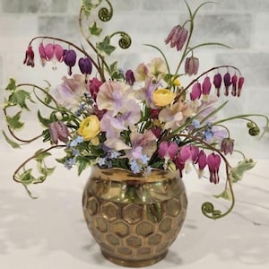 May include: A metallic gold vase with a textured honeycomb pattern holds a floral arrangement featuring purple bleeding hearts, yellow ranunculus, pale lavender sweet peas, small blue forget-me-nots, checkered fritillaria, and green ivy vines.
