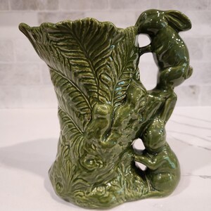 May include: A glossy dark green ceramic pitcher featuring a textured leaf pattern on the body and a handle formed by two sculpted rabbits climbing vertically. The vessel has a wide, flared rim and a detailed, embossed surface throughout the exterior.