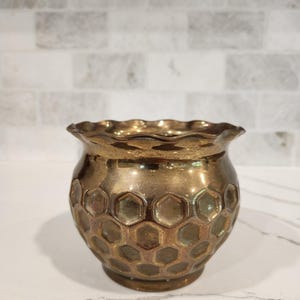 May include: A small, round, metallic bronze-toned vase featuring a raised honeycomb pattern across its exterior surface. The vessel has a wide, scalloped, and ruffled rim, with a smooth, circular base that tapers inward toward the bottom.