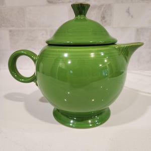 May include: A vibrant green ceramic teapot with a matching lid. The teapot has a rounded body, a curved handle, and a spout. The lid is topped with a small, conical finial. The teapot sits on a pedestal base. The glossy finish reflects light.