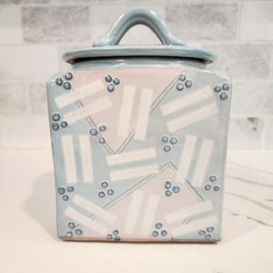 May include: Square ceramic jar with a lid and handle. The jar is light blue with a geometric pattern of white rectangles and pink triangles. Small blue dots are scattered across the surface.