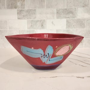 May include: A red ceramic bowl with a square shape and angled sides. The bowl is decorated with blue and pink abstract brushstrokes. The bowl is sitting on a white surface.