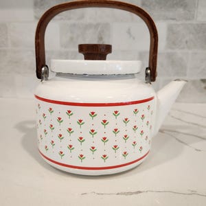 May include: White enamel tea kettle with a wooden handle and lid knob. The kettle features a red and green floral pattern around the body and a red stripe at the base. The handle is arched and made of wood. The spout is on the right side.