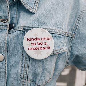 May include: A circular pinback button featuring a white background with light gray star patterns and the maroon text "kinda chic to be a razorback" pinned to a light blue denim jacket with visible stitching and metal buttons.