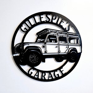 Może przedstawiać: Czarny metalowy znak z tekstem "Gillespies Garage" w kółku. W środku kółka znajduje się biała sylwetka Land Rovera z bagażnikiem dachowym.