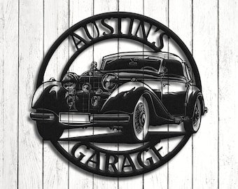 Op de afbeelding: Zwart metalen bord met een vintage auto ontwerp. Het ronde bord heeft de woorden "AUSTIN'S GARAGE" uitgesneden rond de auto. De auto is een klassiek model met gedetailleerde kenmerken. Op een witte houten achtergrond.