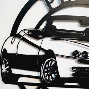 May include: Black and white metal silhouette of a convertible car. The car is a side profile with the front wheel visible. The car is part of a larger design, with a circle around it.