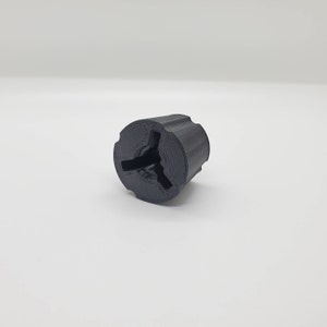 May include: A matte black, cylindrical 3D-printed component featuring a three-pronged internal structure and a textured exterior surface. The object has a circular top face with a recessed center and a solid, uniform dark finish throughout its design.