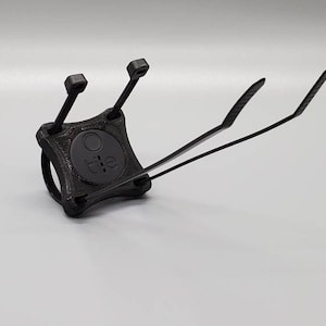 May include: A black 3D-printed mounting bracket designed to hold a circular Tile tracker. The square-shaped mount features four integrated slots for zip ties, with two black zip ties currently threaded through the corners of the device.