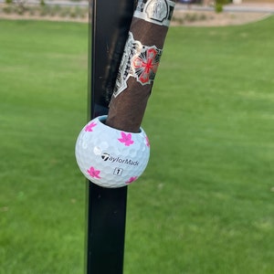 May include: A white golf ball with pink cherry blossom designs and the text "TaylorMade" and "1" on it. The golf ball is attached to a black pole.