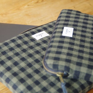 Peut inclure: Deux housses pour ordinateur portable en tissu écossais vert et noir avec fermetures éclair. La plus grande housse a une étiquette blanche avec le texte "HEY MATE" et un petit logo d'oiseau. La plus petite housse a une étiquette blanche avec le texte "STOCKHOLM" et un petit logo d'oiseau.