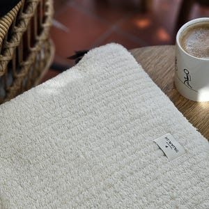 Peut inclure: Une serviette moelleuse blanche avec une petite étiquette blanche sur laquelle est inscrit "HUMMING BIRD". La serviette est posée sur une table en bois.