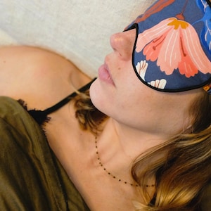 May include: A woman wearing a floral sleep mask with a blue background and pink flowers. She is lying on a white couch with a green blanket.