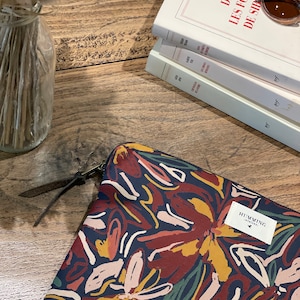 Puede incluir: Una funda para portátil negra con un estampado floral abstracto de colores. La funda tiene una cremallera y un pequeño logotipo que dice "Humming" con un icono de pájaro.