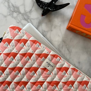 Peut inclure: Une housse d'ordinateur portable matelassée avec un motif d'oiseaux répété en corail, rose et blanc, avec une fermeture éclair dorée. Une figurine d'hirondelle en céramique noire et un livre orange sont également visibles.