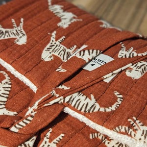 May include: Close-up of a rust-colored, quilted fabric with a repeating pattern of white tigers. The fabric is folded, revealing a white, textured edge. A small tag is visible.