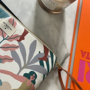 Può includere: Una pochette bianca con cerniera, con un motivo floreale e un tiretto in pelle marrone. La pochette ha il testo "HUMMING" e "atelier." stampato su di essa. La pochette è appoggiata su una superficie di marmo bianco accanto a un libro e una candela.