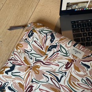 May include: A white laptop sleeve with a floral pattern in shades of brown, pink, green, and black. The sleeve has a zipper closure and a brown leather pull tab. The brand name "HUMMING" is printed on a small tag attached to the sleeve.